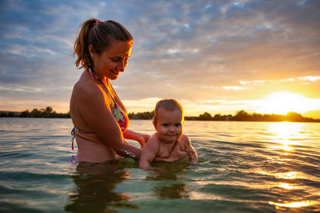 Woman teaching baby swimming in sea during sundown.の写真素材
