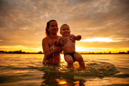 Smiling mother playing holding baby above sea water during sunset.の写真素材