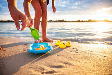 Side crop view of unrecognizable caucasian girl playing with rubber yellow ducks in small blue pool, standing on beach sand. Incognito kid having fun, enjoying holidays at seashore, summer sunset.の写真素材