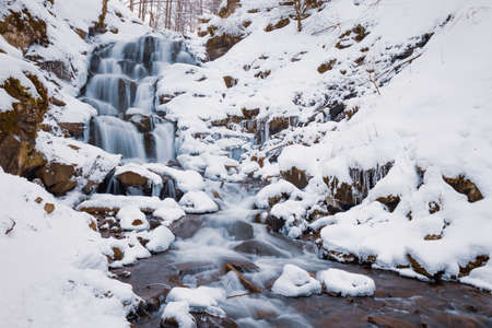 Small mountain waterfall of icy water flow among wet stones covered with white snowの写真素材