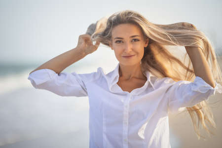 A girl with long hair in a bluish swimsuit and a shirt goes through her hair on the seashoreの写真素材