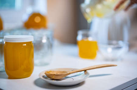 An unknown person pours sweet golden honey into transparent empty clean jars standing on a wooden white large table on a bright sunny dayの写真素材