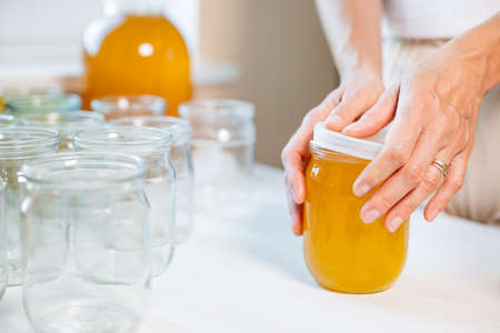 Hands of an unknown woman close transparent glass jars with pure sweet golden honey with a rubber lid, standing on a large white wooden table and next to a small saucer with a wooden spoonの写真素材