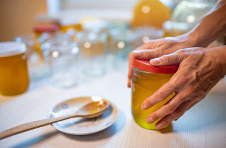 Hands of an unknown woman close transparent glass jars with pure sweet golden honey with a rubber lid, standing on a large white wooden table and next to a small saucer with a wooden spoonの写真素材