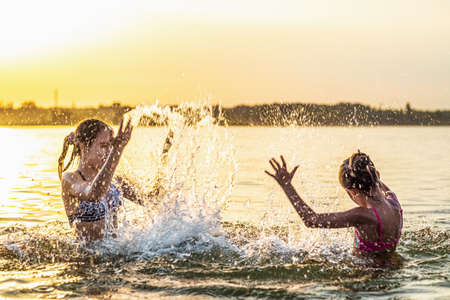 Two funny funny sisters splash on each other with shiny transparent clear water in a blue shallow lake, against the backdrop of a bright sunny golden summer warm sunsetの写真素材