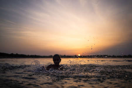 A girl plays with water in the lake and splashes it to the sides against the backdrop of a sunsetの写真素材