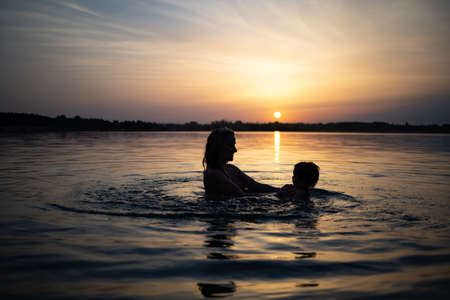Mom plays with a naked baby in oversleeves in the lake against the background of a summer sunsetの写真素材