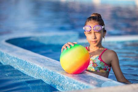 A girl in a bright bathing suit swims with an inflatable ball in a pool with clear water on a summer eveningの写真素材