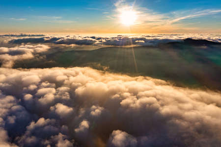 Sun shining brightly on vivid blue sky above thick layer of white fluffy clouds at sunset with mountains on backdrop aerial viewの写真素材