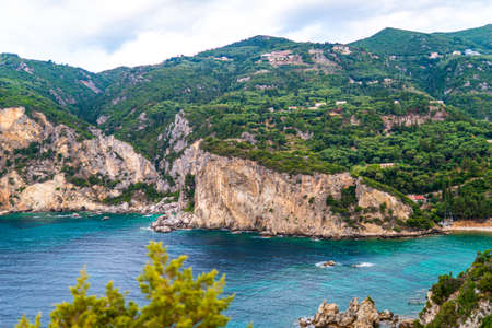 Town buildings standing on high hill top covered with vibrant greenery above tranquil Mediterranean sea blue water under clear skyの写真素材