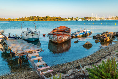 Old shabby wooden row boat moored with long ropes at stones against pier with big yachts of Corfu island in Greece at sunset panoramic viewの写真素材