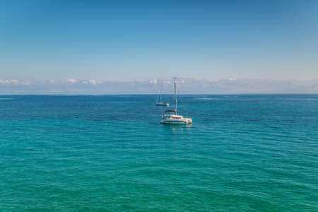 Anchored motorboat on calm water surface against Corfu island with buildings and mountains under blue sky with white puffy cloudsの写真素材