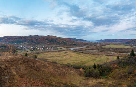 Long railroad bridge over narrow shallow river leading to mountain settlement in valley surrounded by terracotta autumn forests and fieldsの写真素材