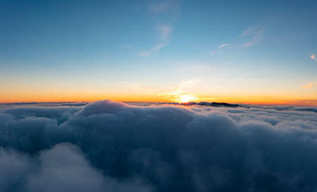 Blue sky above thick layer of white fluffy clouds at bright orange-colored sunrise with mountain peaks underneath panorama viewの写真素材