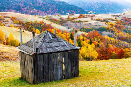 Small wooden haystack house built on grassy slope in highland near colorful trees growing against forestry mountains on bright autumn dayの写真素材