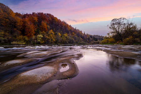 Shallow mountain river rapids with evergreen autumn terracotta forests on steep banks, long exposure...