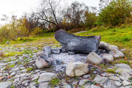 Wooden stump lies on rocks on river stone dried bottom against high cliff with lush terracotta and yellow trees under cloudy sky in autumnの写真素材