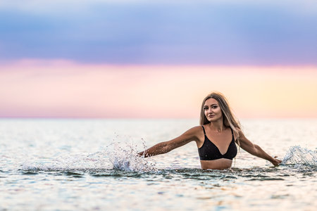 A girl with blond hair in a black swimsuit splashes to the sides while sitting in an estuary on a sunset backgroundの写真素材