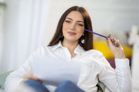 An adult attractive dark-haired girl in a neat stylish white shirt sits near a desk and draws her...