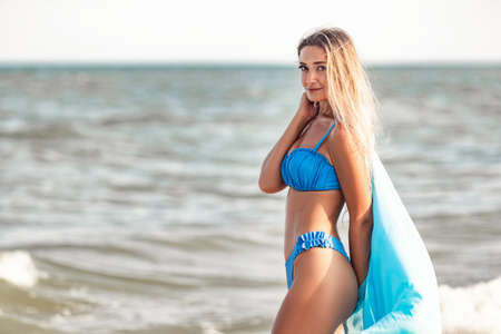 A girl with blond hair in a bluish swimsuit and a bright shawl walks along the beach