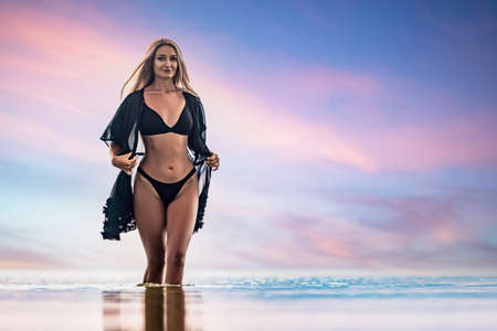 A girl with blond chic hair in a black bikini and a lace shawl walks along the estuaryの写真素材