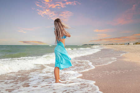 A girl with blond hair in a bluish swimsuit and a bright shawl walks along the beachの写真素材