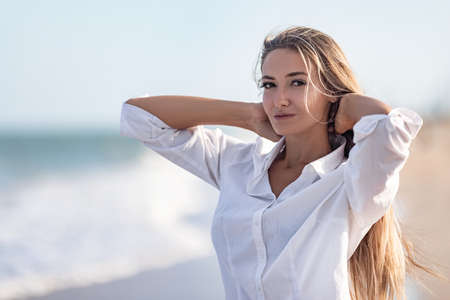 A girl with long hair in a bluish swimsuit and a shirt goes through her hair on the seashoreの写真素材
