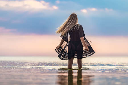 A girl with blond chic hair in a black bikini and a lace shawl walks along the estuaryの写真素材