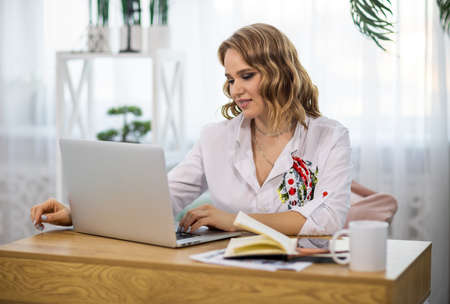 Blonde interested girl in stylish shirt with embroidery sits at desk with her work things and cup of...