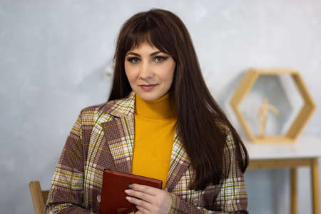 Stylish fashionable dark-haired female model in brown and beige trouser suit and mustard high-neck golf notepad in her hands, against background of dressing table and gray wall in spacious studioの写真素材