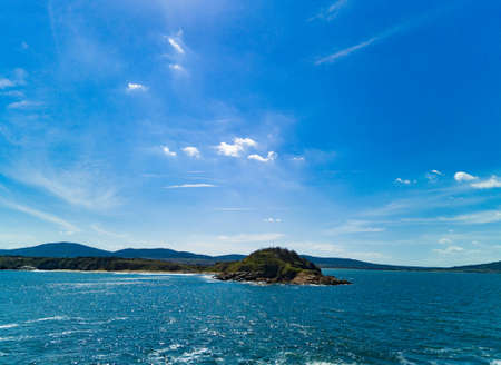 Panorama of a wild stone and sandy beach with dark wet sea sand and large wet rocky stones near the reflective undulating spring Black Sea, under bright sunny sunset light in Bulgariaの写真素材