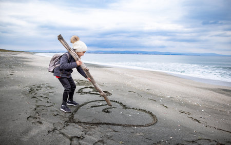 A little dreamy dark-haired girl in warm clothes and a white hat with a large hiking backpack walks on my empty sea sandy beach, and draws childrens drawings and symbols on the wet sandの写真素材