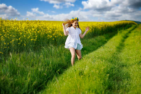 Teenage girl in white dress and Ukrainian wreath runs through yellow fields and green meadows, against cloudy skyの写真素材