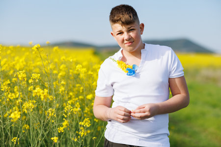 A big kind strong teenage guy in a white T-shirt with field Ukrainian wild flowers on it enjoys the warm summer bright sun while standing, in a yellow blooming rapeseed field against a blue clear skyの写真素材