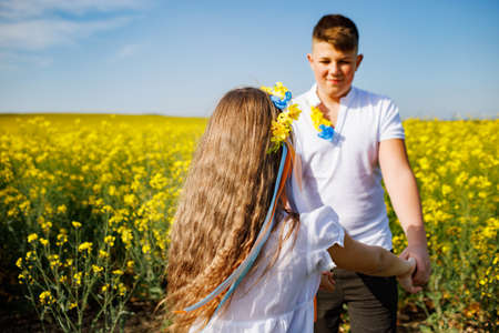 Cheerful joyful teenagers: older brother and laughing sister with floral Ukrainian bright wreath with multi-colored ribbons on head, fools around in yellow rapeseed blooming field under clear blue skyの写真素材