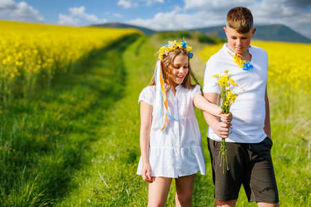 Front view of happy carefree native children: older teenage brother and younger sister walking far away along path with green grass surrounded by yellow blooming rapeseed fields against cloudy skyの写真素材