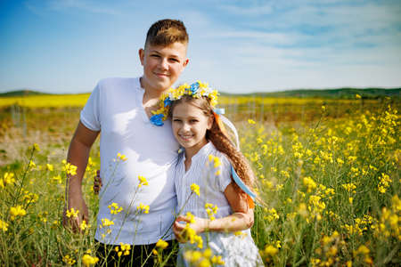 Kind teenage brother carefully hugs cheerful laughing sister with Ukrainian flower wreath with ribbons and smelling neat bouquet of flowers in hands, in yellow rapeseed blooming field under clear skyの写真素材