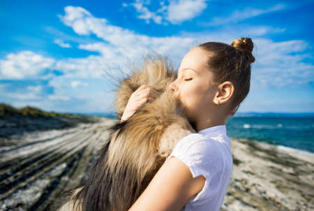 Girl with neatly braided blond hair with cheerful smile in white T-shirt hugs and kisses her fluffy...