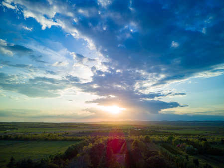 Multi-colored boundless clear sky with white fluffy mighty clouds and a bright golden radiant sun, over green planted farm fields and cattle-breeding small grassy pasturesの写真素材