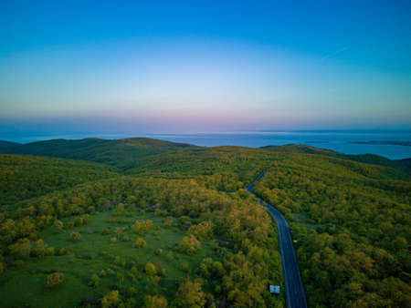 A winding new mountain road with fast modern cars and trucks leads into the distance, beyond the horizon, between green deciduous forests on the hills and the Balkan Mountainsの写真素材