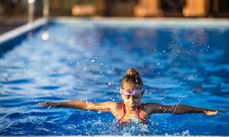 A little girl in a bright pink swimsuit with transparent black goggles for swimming, straightens her goggles and then dives into a deep blue pool with a chimta of clear waterの写真素材