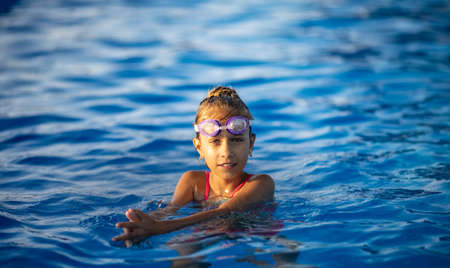 A little girl in a bright pink swimsuit with transparent black goggles for swimming, straightens her goggles and then dives into a deep blue pool with a chimta of clear waterの写真素材