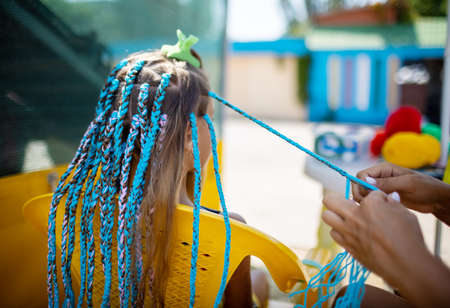 A little waiting tanned girl in a bright yellow summer suit, weaves colored pink-blue African braids in her dark wet thick hair on a hot sunny sea dayの写真素材