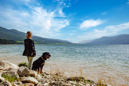 Little girl stands near her big dog friend of Rottweiler breed on empty wild rocky shore near transparent turquoise lake, against backdrop of green Rhodope mountain range covered with spruce forestの写真素材
