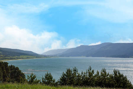 A gleaming wide clear lake reflects bright warm sunlight, against a clear blue cloudy sky and...