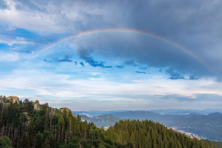 Bright multicolored natural rainbow with different colors on a light daytime cloudy sky, over a high forested mountain valley of the Rhodope Mountains rocks and hillsの写真素材