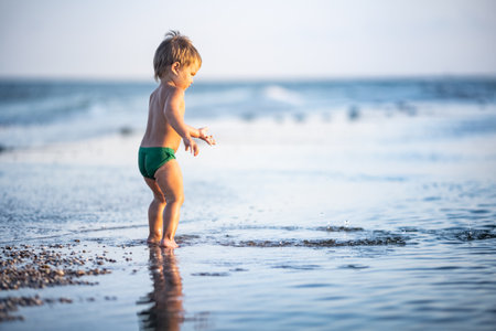 Little funny funny kid, collects shells and pebbles in the calm blue sea on a sandy bottom under the hot summer sun on a bright vacationの写真素材