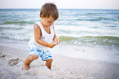 Little cute cheerful kid in a striped gray-white T-shirt looks for and collects wet pebbles and...