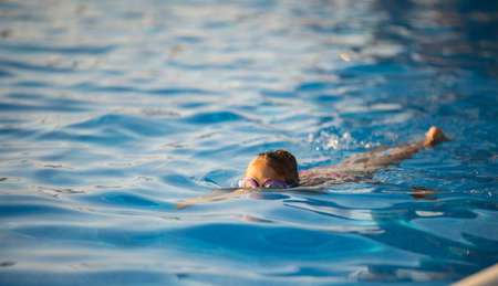 A little girl in a bright pink swimsuit with transparent black goggles for swimming, straightens her...