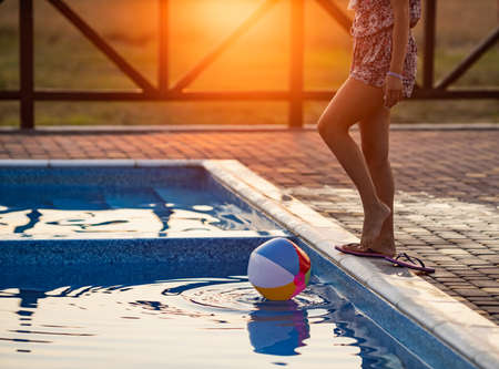 A girl with hair braided in a bun in bright suit plays by the pool with a ball against the background of summer sunの写真素材